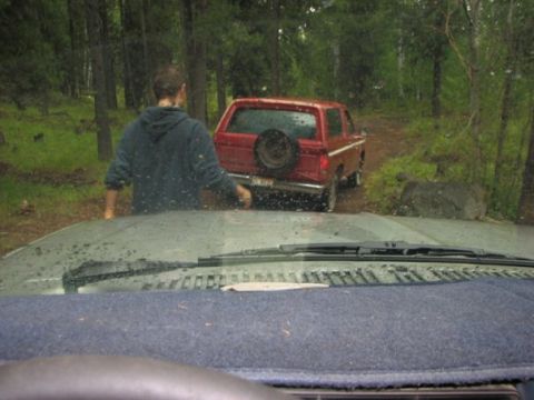 1981 Ford Bronco - A red 1981 Ford Bronco is seen from the perspective of another vehicle, parked on a dirt road in a wooded area.