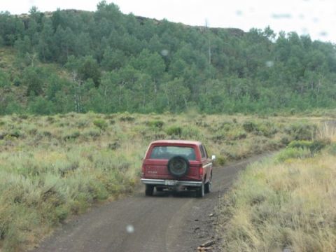 1981 Ford Bronco - Red SUV drives down a dusty road surrounded by dry grass and trees.