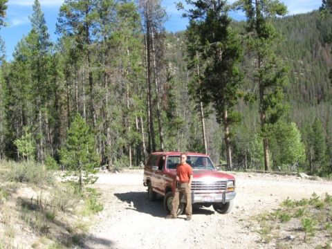 1981 Ford Bronco - A person stands next to a red SUV on a dirt road surrounded by trees and mountains.