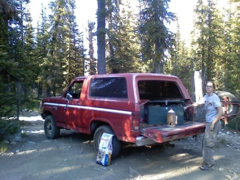 1981 Ford Bronco - A red Ford Bronco is parked in a wooded area with its tailgate open, revealing camping equipment.