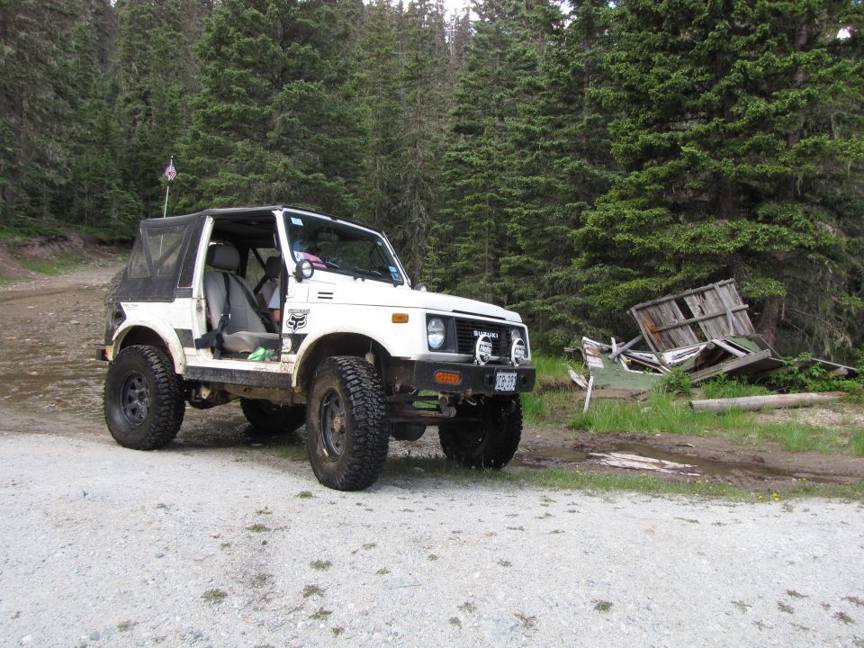 White Suzuki Samurai with doors off and big tires parked by a collapsed cabin in pines