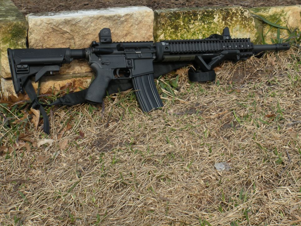 AR-15 rifle with red dot sight and foregrip resting on grass and rocks