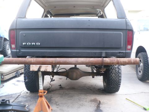 1979 Ford Bronco - The rear of a Ford Bronco is shown, supported by jack stands with a large pipe across the frame.