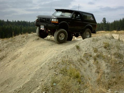 1995 Ford Bronco - A modified black Ford Bronco navigates a dirt incline under a cloudy sky.