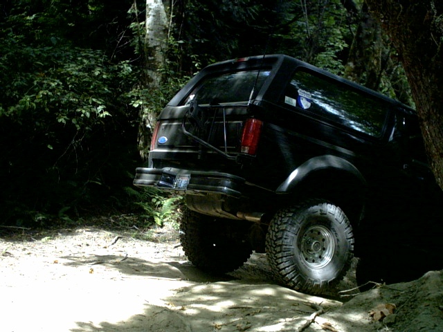 Black Ford Bronco SUV climbing rocks on a shaded forest off-road trail, rear three-quarter view