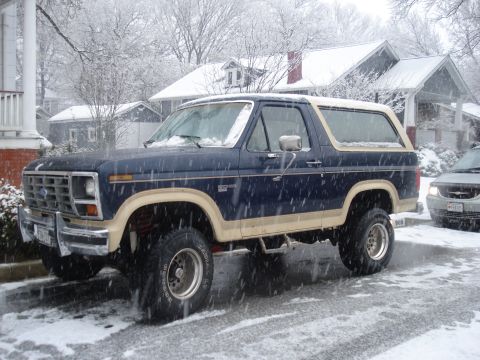 1985 Ford Bronco - A lifted Ford Bronco sits on a snow-covered street in front of houses.