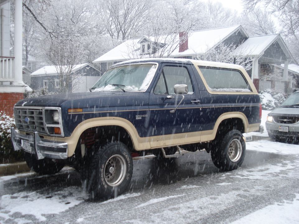 Dark blue Ford Bronco with tan trim, lifted suspension, and aftermarket wheels parked on a snowy street