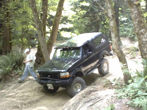 1995 Ford Bronco - A black Ford Bronco navigates a wooded trail with a person walking nearby.