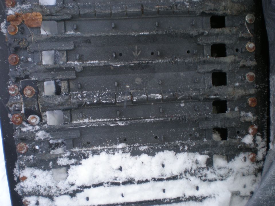 Snow-dusted rubber track with molded lugs, drive holes, directional arrow, and rusty bolts along edges