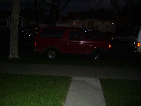 1988 Ford Bronco - Dark red full-size Ford Bronco parked at the curb of a residential street at night, photographed in profile from across a lawn and sidewalk…