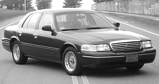 1998 Ford Crown Victoria - A dark 1998 Ford Crown Victoria sedan is shown from the front-right quarter angle while driving on a two-lane road.