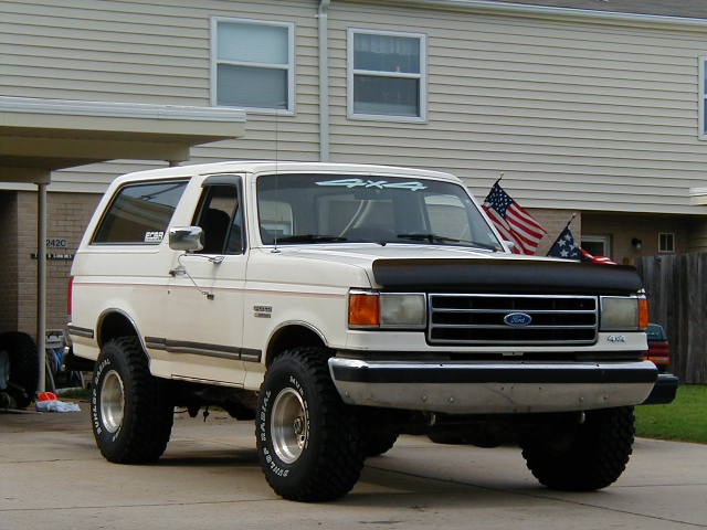 White two-tone lifted Ford Bronco with hood bra and mud tires in a driveway
