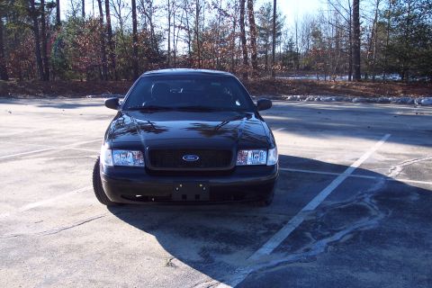 2001 Ford Crown Victoria - A black 2001 Ford Crown Victoria is parked in a lot with trees in the background.