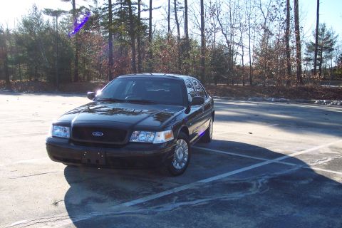 2001 Ford Crown Victoria - A black 2001 Ford Crown Victoria is parked in an outdoor lot with trees in the background.