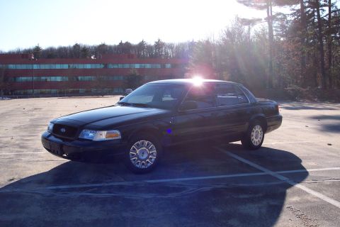 2001 Ford Crown Victoria - A black sedan is parked in an empty lot with a building and trees in the background.