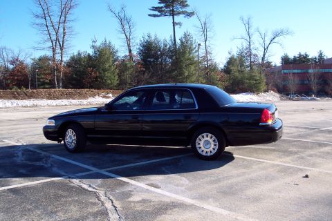 2001 Ford Crown Victoria - Side view of a black 2001 Ford Crown Victoria parked in an empty lot with trees in the background.