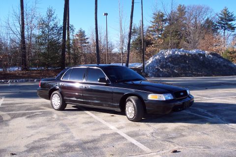 2001 Ford Crown Victoria - A black sedan is parked in an empty lot on a sunny day with trees and a large snow pile behind it.