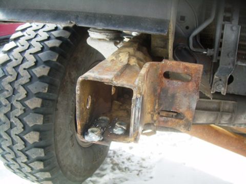 1995 Ford Bronco - Underbody close-up of the rusty front frame horn on a truck, showing the boxed frame end, a welded bumper mounting bracket with slotted hol…