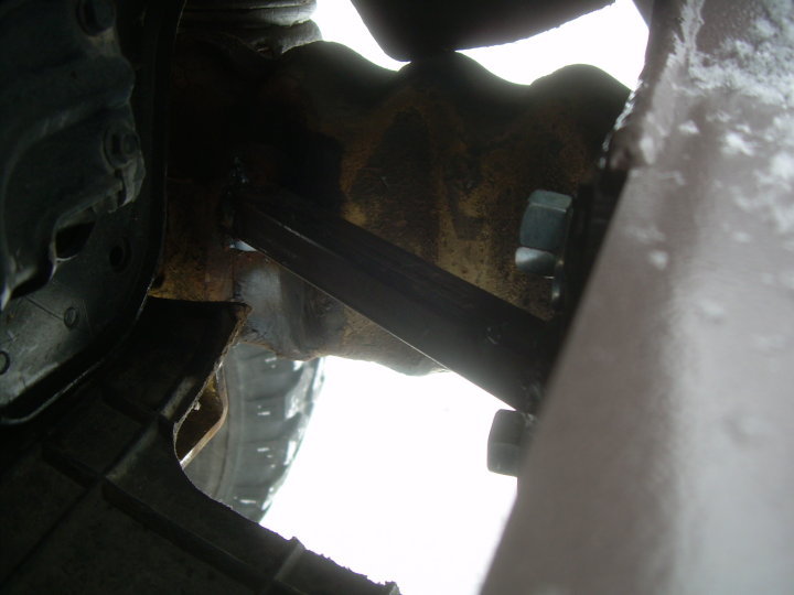 Rusty truck frame rail with black brace and hex bolts behind a gray bumper