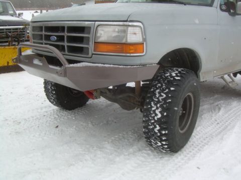 1995 Ford Bronco - Close front three-quarter shot of a light gray 1990s Ford truck in falling snow, showing a shop-built flat plate steel front bumper in bare…