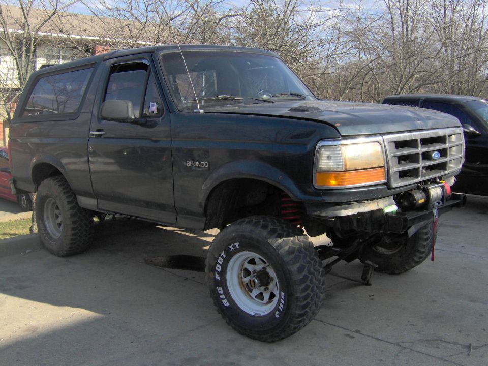Lifted dark green Ford Bronco with white wheels, Big Foot XT tires, and front winch, parked on concrete driveway