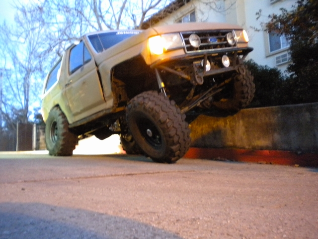 Lifted tan Ford Bronco flexing a front wheel up a concrete wall at dusk