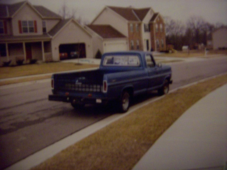 Blue Ford F-100 pickup parked on a suburban street, seen from the rear three-quarter angle