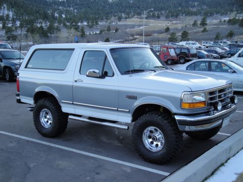 1995 Ford Bronco - This lifted Ford Bronco is parked in a lot with other vehicles and a mountain range in the background.