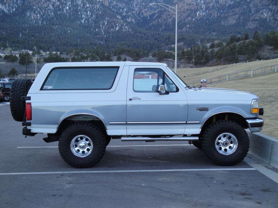 Silver two-tone Ford Bronco parked on asphalt, side view, spare tire on rear door