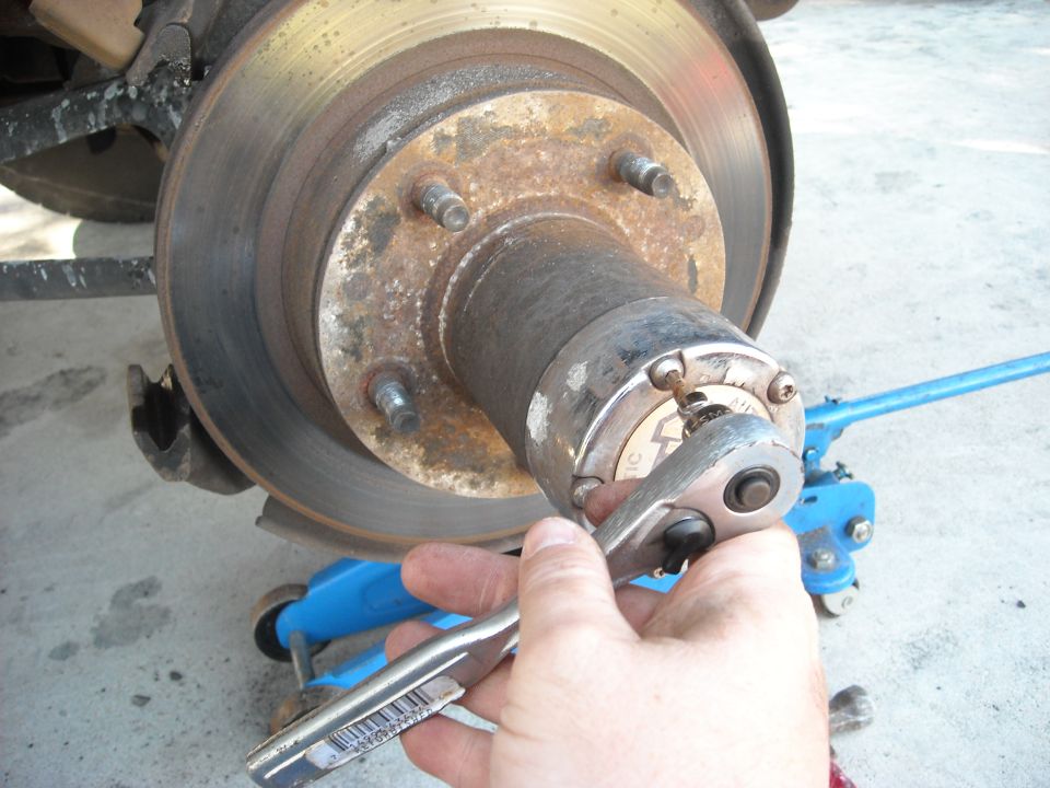 Close-up of a vehicle's front wheel hub assembly with a socket wrench being used on a nut. A blue jack is visible in the background