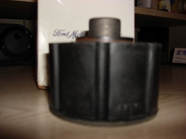 Close-up of a black cylindrical part with ribbed sides, sitting on a wooden surface. A white box with 'Ford Motor' visible is behind it