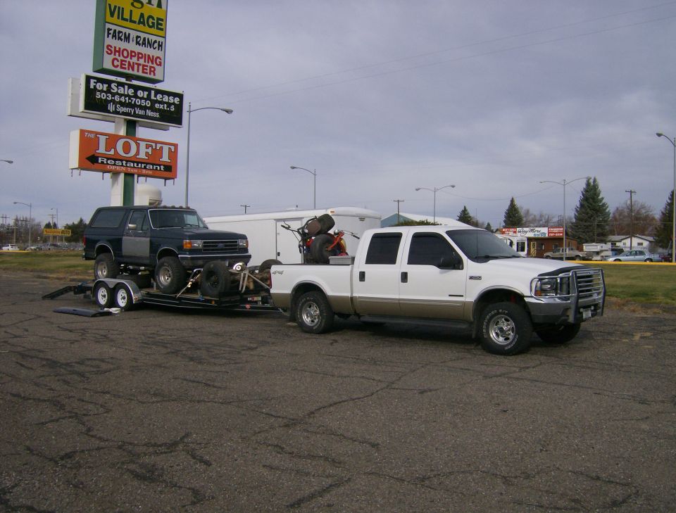 White 2000 Ford F-250 towing a trailer with a dark-colored Bronco on it. Signage for 'The Loft Restaurant' and 'Village Farm Ranch Shopping Center' is visible