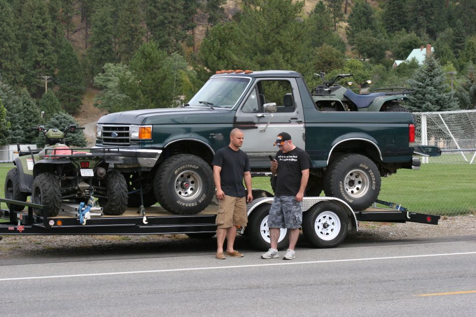 Two guys standing by a dark green Ford Bronco on a trailer, with two ATVs also on the trailer