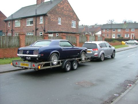 1970 Ford Mustang - A blue 1969 Ford Mustang coupe is transported on a trailer, towed by a silver SUV on a residential street.