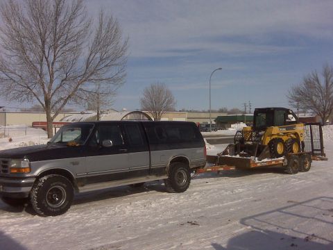 1994 Ford F-150 - A Ford pickup truck tows a skid steer loader on a trailer through a snowy landscape.
