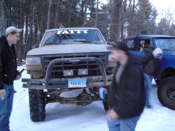 Muddy lifted pickup with 'FATTY' on windshield in snow, blue SUV behind