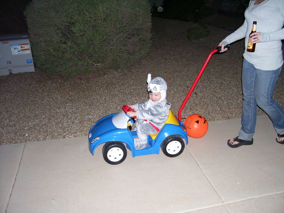 Kid in a blue toy car with a push handle, being pushed by an adult holding a beer