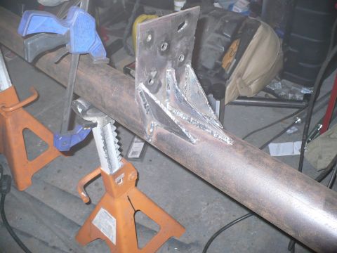 1979 Ford Bronco - A metal bracket is welded onto a pipe supported by orange jack stands in a workshop.