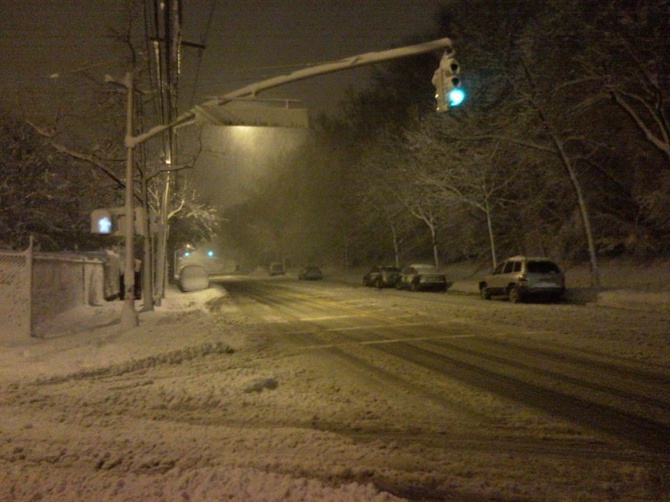 Snowy street at night with traffic lights and several cars parked or driving