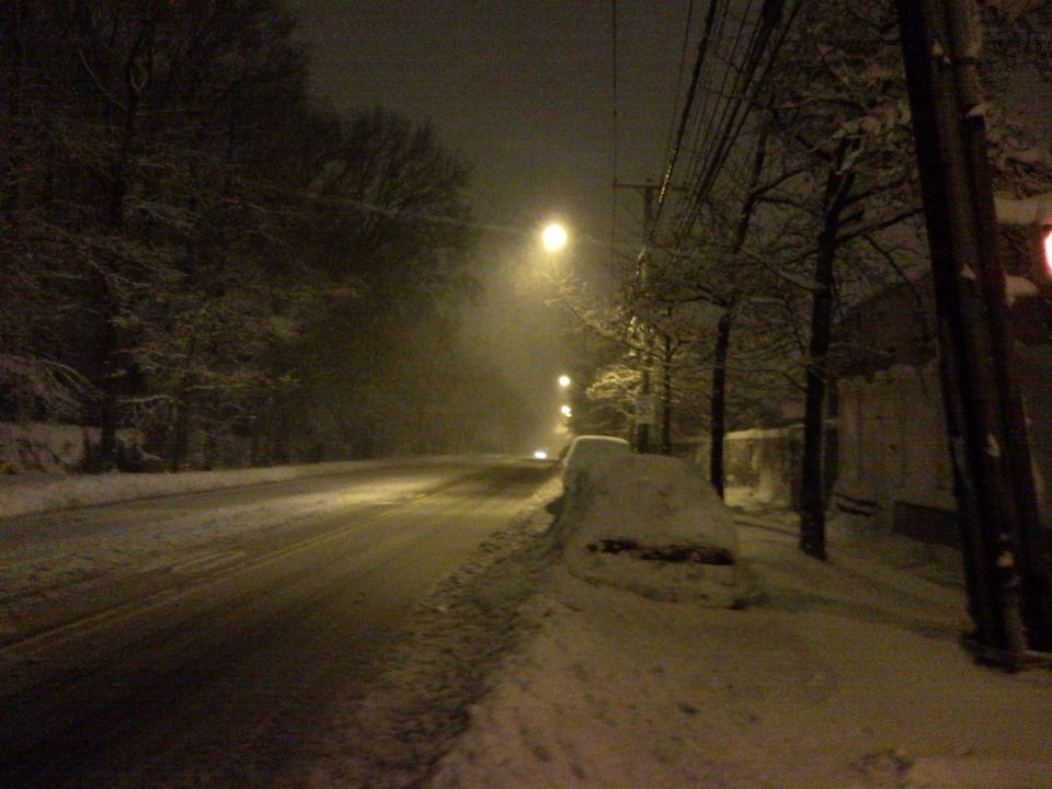 Streetlights illuminate a snow-covered road at night with trees and a partially buried car on the side