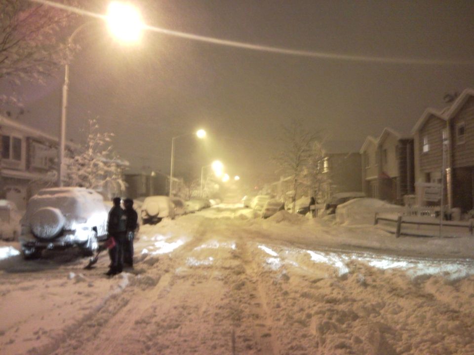 Street buried in snow with parked cars and two people shoveling