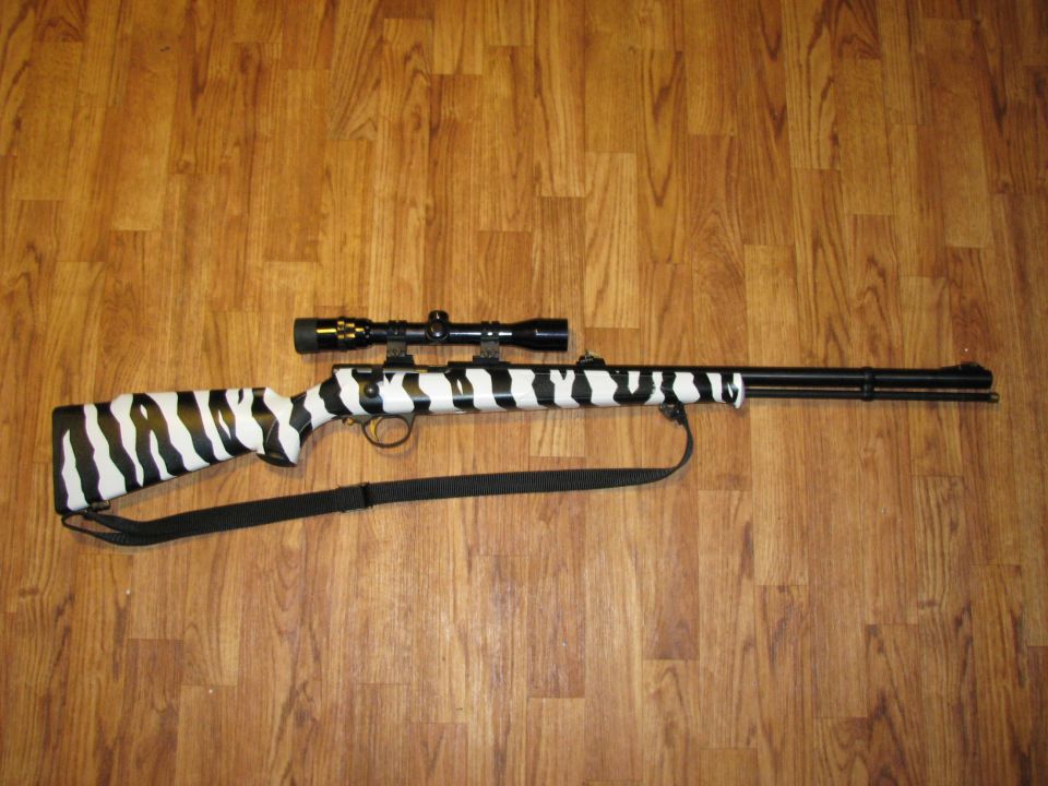 Rifle with black and white zebra pattern, mounted scope, and black strap on wooden floor