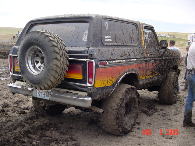 Mud-covered full-size Bronco with sunset-stripe graphics and rear spare, stuck axle-deep in a mud pit