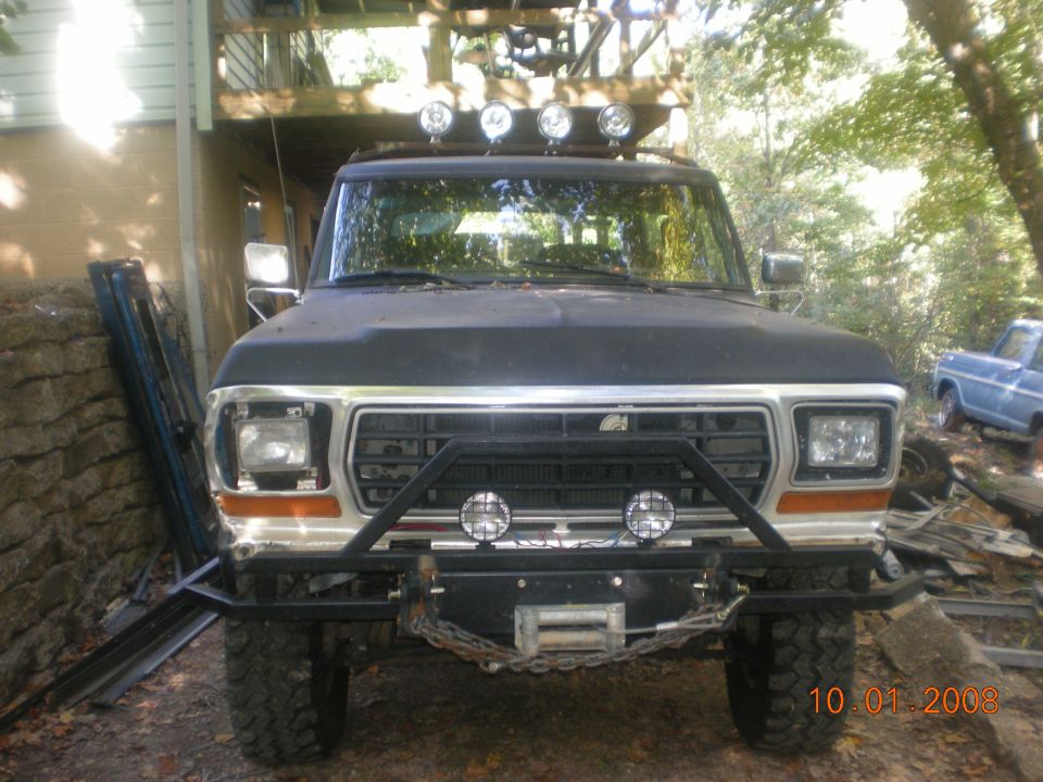 Black Ford Bronco with winch and roof lights, front view. A blue pickup truck is visible in the background