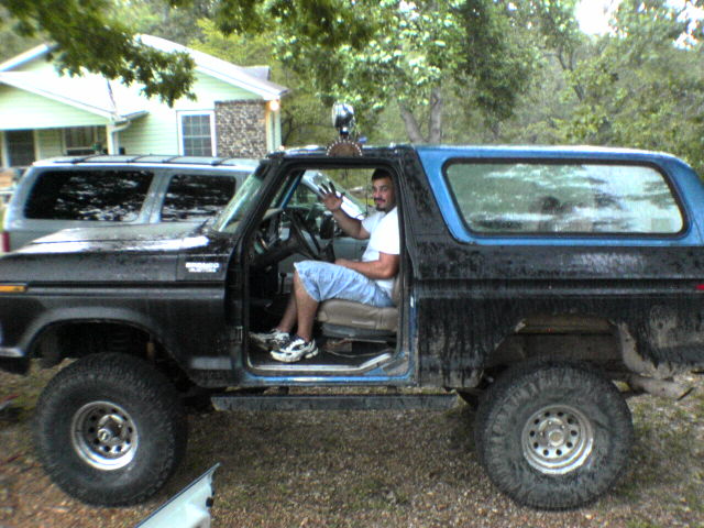 Man in driver's seat of a black Ford Bronco with a blue top. Another SUV is parked behind it