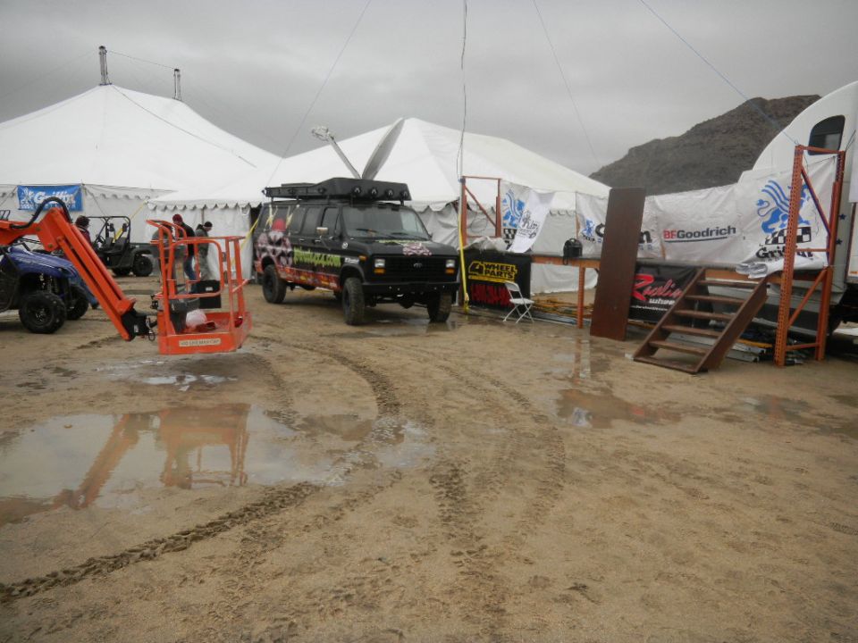 Black van with roof rack and off-road tires parked near tents. Orange lift and blue ATV also visible