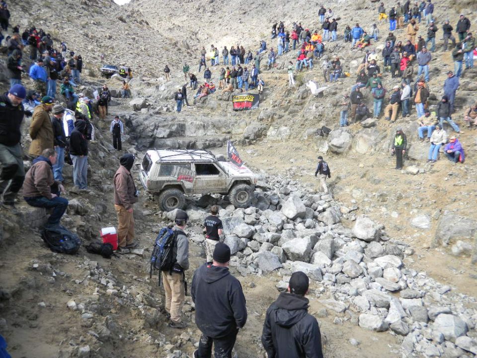 White off-road SUV with a roof rack and flag on a rocky slope, spectators watching