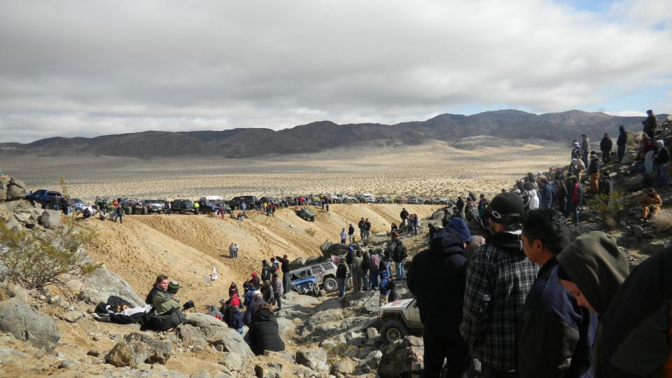 Off-road vehicles and spectators gathered in a desert terrain with hills and mountains