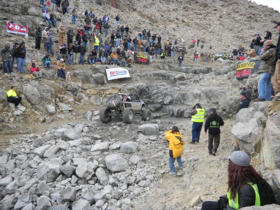 Off-road buggy with spare tire on back, climbing rocks with spectators watching