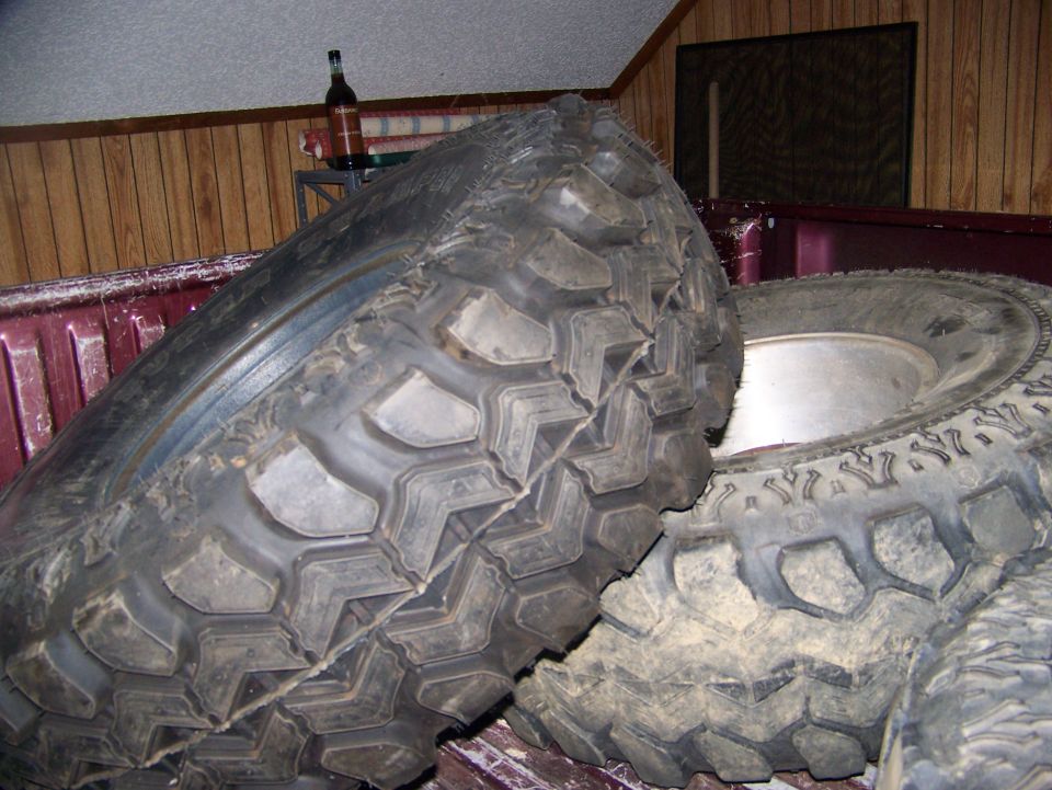 Two large, knobby off-road tires piled in the bed of a dark red pickup truck